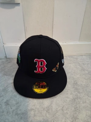 Svart Boston Red Sox keps New Era - Svart keps från New Era med Boston Red Sox-logga framtill i rött och vitt. Kepsen har broderade fjärilar och blommor på sidan samt klassisk flat skärm. Perfekt accessoar för dig som gillar sport och streetstyle.