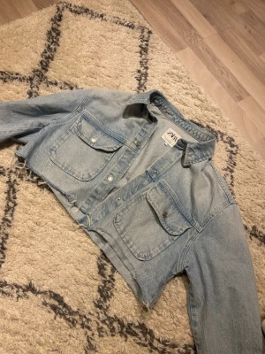 Cropped ljusblå jeansjacka från Zara - Trendig cropped jeansjacka från Zara i ljusblå denim. Jackan har två stora bröstfickor med knappar. Perfekt att style med jeans eller klänning 🩵