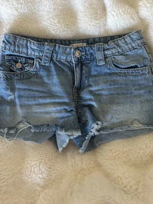Blå jeansshorts med fransig kant - Säljer ett par blå jeansshorts med fransig, rå kant och slitna detaljer. Shortsen har fem fickor, bälteshällor och knappgylf. Perfekta för varma dagar och ger en avslappnad look.