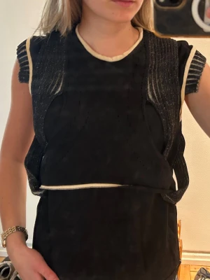 Svart Dagmar topp - Trendig svart stickad topp med vita kontrastsömmar och råa kanter. Toppen har korta ärmar och en unik, strukturerad design med insydda partier och markerad midja. Perfekt för dig som vill sticka ut med en cool och modern look.