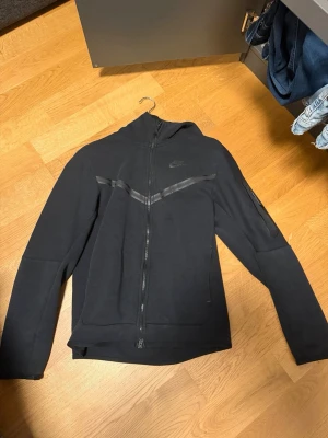 Svart Nike Tech Fleece hoodie (finns byxor) - Går att köpa med byxor!!! Svart Nike Tech Fleece hoodie med hel dragkedja och huva. Snygga svarta detaljer i form av två diagonala ränder över bröstet och diskret Nike-logga på vänster sida. Mjuk och lätt fleece som passar perfekt till streetwear-stilen.