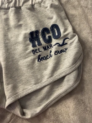 Grå hollister shorts - Snygga ljusgrå shorts från Hollister med broderad text 'HCO Del Mar beach crew' i mörkblått på ena benet. De har elastisk midja och sportig passform med rundade kanter. Perfekta för sommardagar eller strandhäng.
