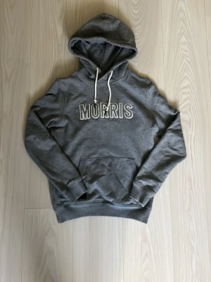 Grå hoodie från Morris med tryck - Snygg grå hoodie från Morris med vit broderad logga på bröstet. Hoodien har huva med vita dragsnören och en klassisk känguruficka framtill. Perfekt för en avslappnad och trendig stil. Mjuk bomullsblandning och ribbade muddar vid ärmslut och nederkant.