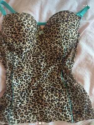 Leopardmönstrad body med turkosa band - Snygg body med leopardmönster och vadderade kupor. Turkosa axelband och dekorativa sömmar ger en cool kontrast. Materialet är stretchigt och följsamt, perfekt för att framhäva figuren. Liten rosett framtill för extra detalj.