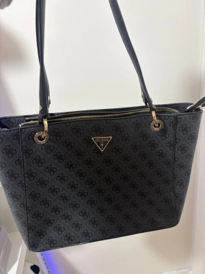 Handväska från Guess - Snygg handväska från Guess med klassiskt logomönster över hela väskan och guldfärgade detaljer. Väskan har dubbla axelremmar och dragkedja upptill. Perfekt för dig som gillar stilrena accessoarer med en lyxig touch.