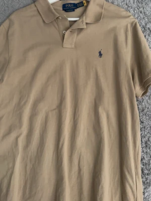 Beige pikétröja från Ralph Lauren - Klassisk beige pikétröja från Ralph Lauren med blå broderad logga på bröstet. Tröjan har krage, korta ärmar och knäppning framtill. Perfekt för en stilren och avslappnad look.