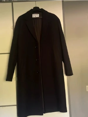 Svart lång kappa från Filippa K - Stilren svart lång kappa från Filippa K med klassisk krage och knäppning framtill. Kappan har rak passform och är tillverkad i ett mjukt tyg som ger en elegant look. Perfekt för dig som gillar minimalistisk och tidlös stil.