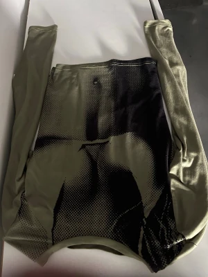Grön långärmad mesh topp med tryck - Cool långärmad mesh topp i olivgrönt med svart grafiskt tryck framtill. Tunn och stretchig med kroppsnära passform, perfekt för att sticka ut. Passar dig som gillar edgy och unik stil.