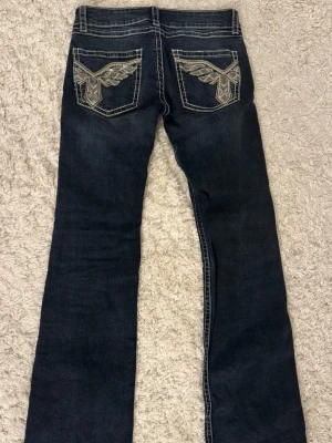 Lågmidjade y2k jeans  - Snygga mörkblå bootcut jeans med kontrastsömmar och broderade bakfickor med vingar. Jeansen har låg midja och klassisk femficksdesign. Perfekta för dig som gillar detaljer och en cool stil.