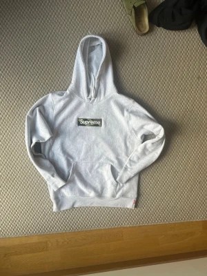 Supreme Hoodie - Supreme hoodie i ljusgrått med klassisk camo box logo på bröstet. Hoodien har huva med dragsko och en stor magficka. Mjuk bomullsblandning och ribbade muddar vid ärmslut och nederkant. Perfekt för dig som gillar streetwear och vill sticka ut.
