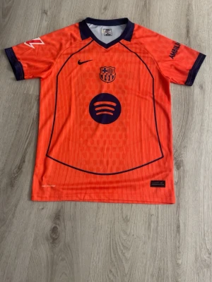 Barça orange matchtröja Nike - Snygg orange FC Barcelona matchtröja från Nike med mörkblå detaljer och klubbmärke på bröstet. Tröjan har korta ärmar, V-ringning och tryckta loggor från Spotify och Ambilight. Perfekt för fotbollsträning eller att bära som supporter.