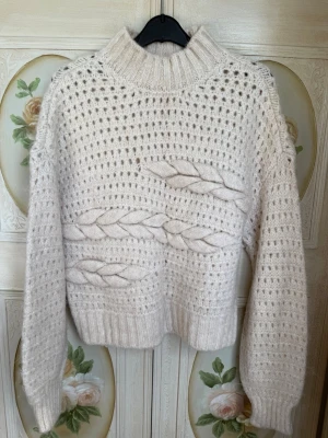 Beige stickad tröja med flätmönster - Enkel och stilren design som passar till det mesta. vit stickad tröja med höghals och snygga flätade detaljer framtill. Tröjan har ett luftigt hålmönster och ribbade muddar vid ärmslut och nederkant. 