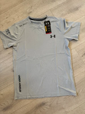 Ljusgrå träningströja från Under Armour - Ljusgrå kortärmad träningströja från Under Armour med svart logga på bröstet och tryck på ärm och nederkant. Tillverkad i ett lätt och snabbtorkande material, perfekt för intensiva träningspass. Rund halsringning och modern passform.