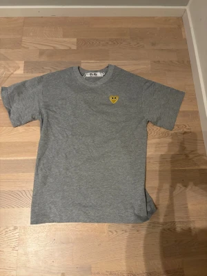 Grå Comme des Garçons PLAY t-shirt - Snygg grå t-shirt från Comme des Garçons PLAY med det ikoniska gula hjärtat med ögon broderat på bröstet. Klassisk rund halsringning och korta ärmar. Perfekt för en stilren och trendig look.