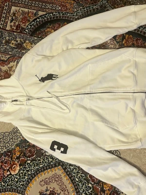 Vit hoodie från Polo Ralph Lauren - Vit hoodie med dragkedja från Polo Ralph Lauren. Stor svart logga på bröstet och siffran 3 på ena ärmen. Klassisk huva med dragsko och två fickor framtill. Skön och stilren, perfekt till jeans eller joggers.