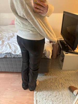 Svarta raka byxor i skinnimitation - Säljer ett par svarta byxor i skinnimitation med rak passform och hög midja. Byxorna har en stilren look och passar perfekt till en trendig outfit. Materialet ger en cool och modern känsla. Perfekta för dig som vill ha ett par statement-jeans.