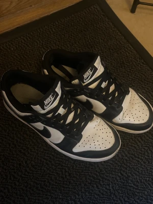 Nike Dunk Low svartvita sneakers - Säljer ett par Nike Dunk Low sneakers i svart och vitt. Skorna har klassisk låg siluett, perforerad tå och Nike-logga på tungan och sidan. Ovandelen är i läder med svarta och vita paneler, och de har svarta skosnören. Perfekta för en streetwear-look.