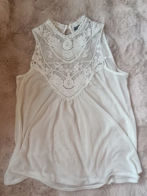Vit blus med spetsdetaljer - Supersöt vit blus med spets och broderade detaljer upptill. Ärmlös modell med rund halsringning och knäppning i nacken. Tunt, lätt material som ger en luftig känsla. Perfekt att styla med jeans eller kjol för en fräsch look. Katt finns i hemmet.