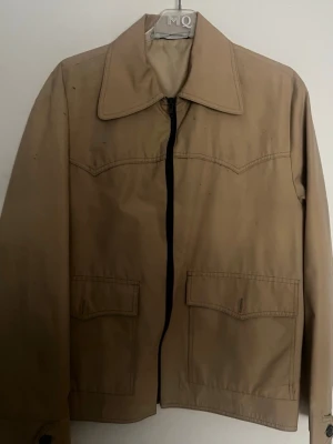Beige jacka med stor krage - Snygg beige jacka från MQ med stor retroinspirerad krage och raka linjer. Jackan har två stora fickor framtill med lock och dragkedja i mitten. Tillverkad i ett slätt tyg, perfekt för dig som gillar minimalistisk stil.
