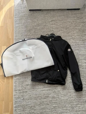 Svart vindjacka från Moncler - Fick den i present så har inget kvitto den är köpt på nordiska kompaniet (NK) i Stockholm Snygg svart vindjacka från Moncler med huva och dragkedja framtill. Jackan har Moncler-logga på ärmen och praktiska fickor med tryckknappar. Tillverkad i ett lätt material som ger en exklusiv känsla. Perfekt för dig som vill ha en stilren och ikonisk jacka. ‼️OBS moncler fodralet till jackan ingår inte‼️jackan är 165-175cm lång borde vara M-L i barnstorlek. Fler bilder går att få. Priset är inte hugget i sten