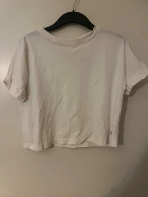 Vit t-shirt i bomull - Enkel och stilren vit croppad t-shirt med rund halsringning och korta ärmar. Tillverkad i mjuk bomull, perfekt för en avslappnad och trendig look. Liten diskret logga nertill på framsidan.