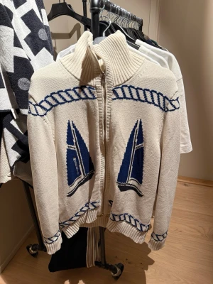 Vit stickad cardigan från Morris - Säljer en vit stickad kofta från Morris med blå segelbåtsmotiv på framsidan. Koftan har dragkedja, hög ribbad krage och blå detaljer längs kanterna. Perfekt för dig som gillar nautisk stil och vill ha något unikt i garderoben.    Ny pris 3999kr