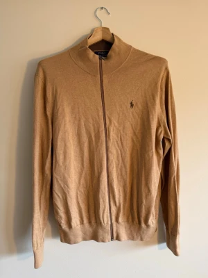 Polo Ralph Lauren - Full zip camel - Polo Ralph Lauren full-zip knit i camel, stl L. Brun dragkedja, mörkbrun ribbekrage inuti – bra detaljer för priset. Tidlös earth tone som funkar som mellanlager eller standalone. Skick: som ny