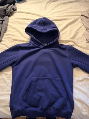 Blå hoodie med stor M-logga - Säljer en blå hoodie från Monki med stor M-logga i tyg på ryggen. Hoodien har klassisk känguruficka framtill och en rymlig huva. Perfekt för dig som gillar enkel men snygg streetstil.