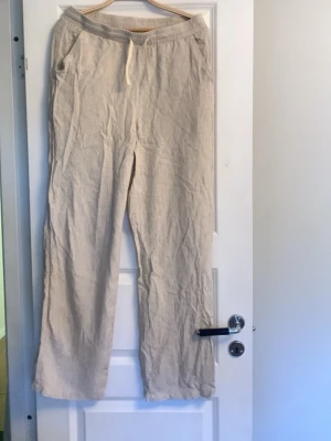 Beiga linnebyxor med dragsko - Snygga och luftiga linnebyxor i beige med raka ben och resår i midjan samt dragsko. Perfekta för en avslappnad stil och sköna varma dagar. Byxorna har fickor framtill och är tillverkade i ett lätt linnematerial.