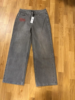 Gråa wide leg jeans från Urban Outfitters i samarbete med Ed Hardy - Snygga gråa jeans från Urban Outfitters i samarbete med Ed Hardy med breda ben och röd broderad logga på både fram- och bakficka. Jeansen har klassisk femficksdesign och är tillverkade i denim. Perfekta för en trendig streetstyle-look.