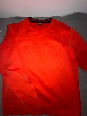 Röd sweatshirt från Hugo Boss - Snygg röd sweatshirt från Hugo Boss med stort BOSS-tryck och broderad logga på bröstet. Tröjan har rund halsringning och är tillverkad i mjukt material som passar perfekt till en avslappnad stil.