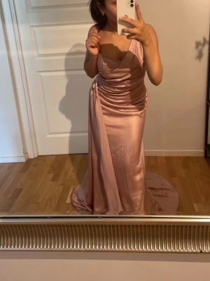 Lång ljusrosa satinklänning - Elegant långklänning i ljusrosa satin med draperad detalj vid midjan och v-ringad överdel. Klänningen har en figurnära passform och ett vackert fallande tyg som ger en lyxig känsla. Perfekt för bal eller festliga tillfällen.