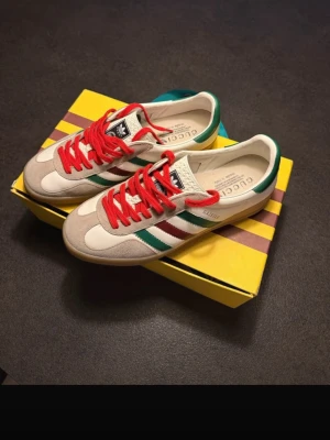 Adidas x Gucci Gazelle sneakers - Snygga Adidas x Gucci Gazelle sneakers i beige mocka och vitt läder med röda skosnören och gröna samt röda ränder på sidorna. Skorna har klassisk låg siluett och detaljer i grönt på hälen. Kommer i exklusiv gul Adidas x Gucci-box.