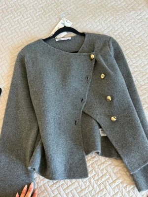 Grå kofta med guldknappar från ZARA - Stilren grå kofta från ZARA med asymmetrisk knäppning och fyra dekorativa guldknappar. Koftan har rund halsringning och är tillverkad i ett mjukt, ulligt material. Perfekt för dig som gillar en clean och modern look.