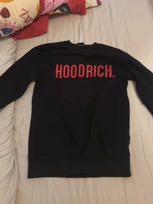Svart sweatshirt från Hoodrich XS - Svart sweatshirt från Hoodrich i storlek XS med stor röd logotyp på bröstet. Tröjan har rund halsringning och långa ärmar. Perfekt för en streetwear-look och enkel att matcha med jeans eller joggers.