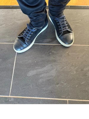 Lanvin mörkblå sneakers med vit sula - Stilrena svarta sneakers med vit sula och mörkblå skosnören. Ovandelen är i en mix av läder och mocka vilket ger en snygg kontrast. Klassisk låg modell som passar till många olika outfits.