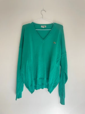 Grön v-ringad tröja från Lacoste - Snygg grön stickad tröja från Lacoste med klassisk v-ringning och den ikoniska krokodilloggan broderad på bröstet. Tröjan har långa ärmar och ribbade muddar vid ärmslut och nederkant. Perfekt för dig som gillar stilrena och tidlösa plagg.