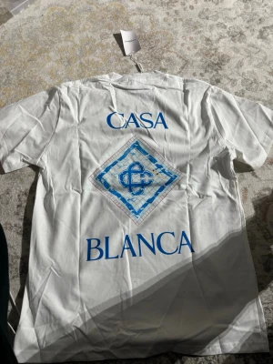 Vit t-shirt från Casablanca - Vit t-shirt från Casablanca med blått tryck både fram och bak. Stort grafiskt motiv och texten 'Casa Blanca' i blått. Klassisk passform med rund hals och korta ärmar. Perfekt för en clean och stilren look.