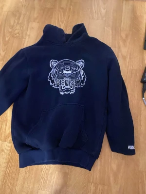 Mörkblå Kenzo hoodie med tigertryck - Snygg mörkblå hoodie från Kenzo med det ikoniska tigerhuvudet broderat på bröstet och Kenzo-logga på ärmen. Hoodien har en stor känguruficka framtill och ribbade muddar. Perfekt för dig som vill sticka ut med stilren streetwear. Tror ej den e äkta derför e priset så lågt 