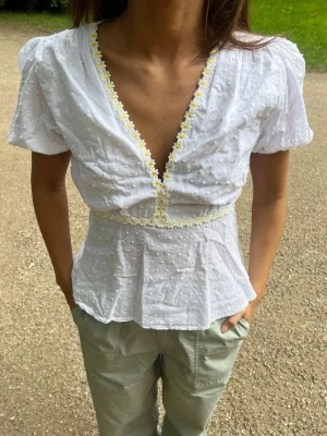 Vit blus med broderade blommor - Söt vit blus med v-ringning och korta puffärmar. Blusen har små broderade prickar i tyget och gula blommor längs urringningen och midjan. Bara använt 1 gång!