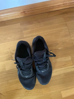 Svarta Nike sneakers mesh och gummi - Säljer ett par helsvarta Nike sneakers med mesh-överdel och gummisula. Skorna har klassisk Nike-logga på sidan och framtill, samt svarta skosnören. Låg modell som passar perfekt till streetwear och vardagsstil.
