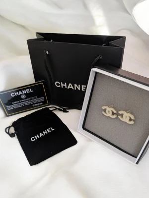 ChaneI -  Design: Elegant sammanflätad logotyp i en sofistikerad ljusguld eller krämvit finish med en silkeslen yta. Tillbehör: Levereras som ett komplett set med originalask, skyddande sammetspåse och matchande papperskasse.
