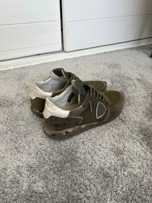 Olivgröna sneakers från Philippe Model - Snygga sneakers från Philippe Model i olivgrönt tyg och mocka med vita detaljer och kamouflagemönstrad sula. Klassisk låg modell med snörning och ikonisk sköldlogga på sidan. Perfekta för dig som gillar streetstyle och unika detaljer.