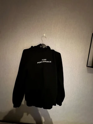 Svart Peak Performance hoodie - Säljer en svart hoodie från Peak Performance med vit logga på bröstet. Klassisk modell med huva och känguruficka framtill. Tillverkad i mjukt och bekvämt material, perfekt för en avslappnad stil. Passar dig som gillar stilrena och sportiga plagg. Detta är en dam hoodie men passar er killar som är runt 170 och väger runt 55-60 annars för tjejer är det som en s 