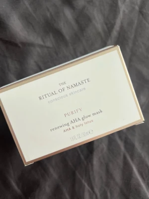 Ritual of Namaste AHA Glow Mask - En ansiktsmask från Ritual of Namaste, Purify renewing AHA glow mask med AHA och helig lotus. Kommer i en vit och guldig rektangulär förpackning, 50 ml. Perfekt för dig som vill ge huden extra lyster och fräsch känsla.