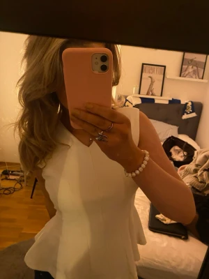 Vit peplumtop med v-ringning - Säljer en snygg vit peplumtop med v-ringning och ärmlös design. Toppen har en figurnära passform upptill och ett volangformat avslut nedtill som ger en cool och feminin look. Perfekt att matcha med jeans eller kjol för en trendig outfit.