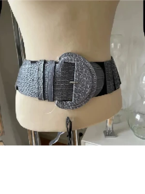 Grått brett bälte med crocoprägling ny  - Snyggt och brett grått bälte med crocopräglat mönster och stor rund spänne. Bältet har elastiska partier på sidorna för extra komfort och passar perfekt till att markera midjan över klänning eller skjorta.