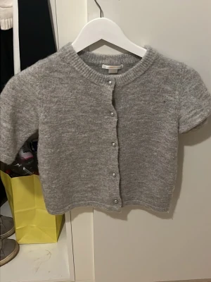 Grå kortärmad kofta med pärlknappar - Söt grå kofta med korta ärmar och pärlknappar framtill. Mjuk stickad känsla och rund halsringning. Perfekt att slänga över en topp för en chill look.