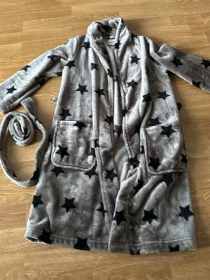 Grå morgonrock med stjärnor Hampton Republic - Mysig grå morgonrock från Hampton Republic med mörkblåa stjärnor, två stora fickor framtill och tillhörande knytskärp. Tillverkad i mjuk fleece som känns skön mot huden. Perfekt för att chilla hemma eller efter duschen.