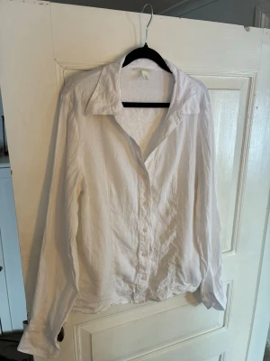 Vit linneskjorta från H&M - Klassisk vit skjorta från H&M i linnemix. Skjortan har lång ärm, knappar framtill och en avslappnad krage. Perfekt till både jeans och kjol för en clean och fräsch look. Passar M/L.